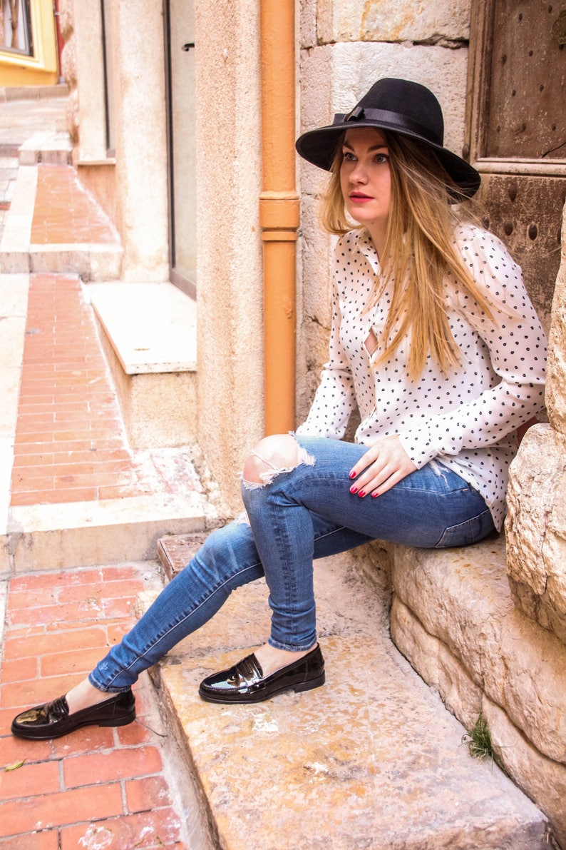 May include: A woman wearing a white polka dot shirt, blue jeans with a distressed knee, and black loafers sits on stone steps. She is wearing a black hat with a black ribbon.