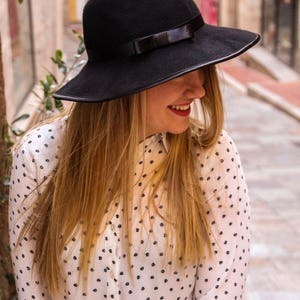 May include: A woman wearing a black fedora with a black ribbon and a white blouse with a black polka dot pattern.