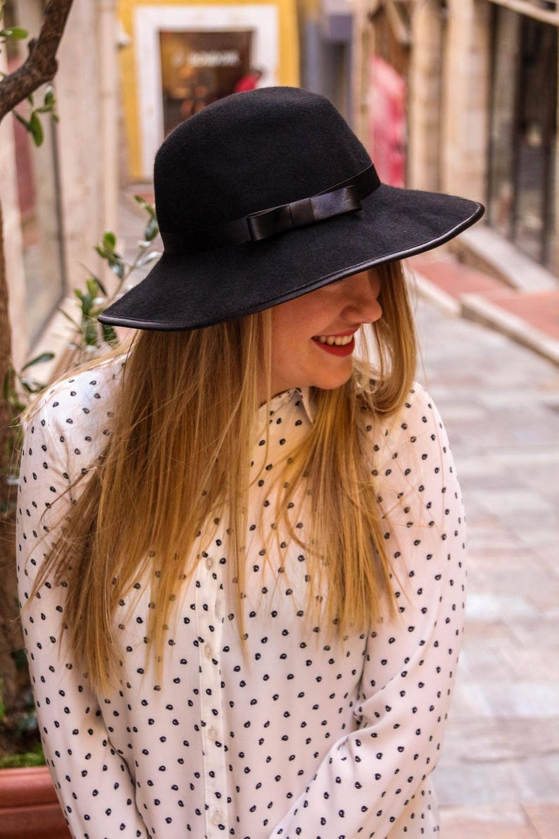 May include: A woman wearing a black fedora with a black ribbon and a white blouse with a black polka dot pattern.