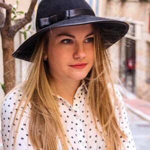 May include: A woman with long blonde hair wearing a black fedora hat with a black ribbon and a white button-down shirt with a black polka dot pattern.