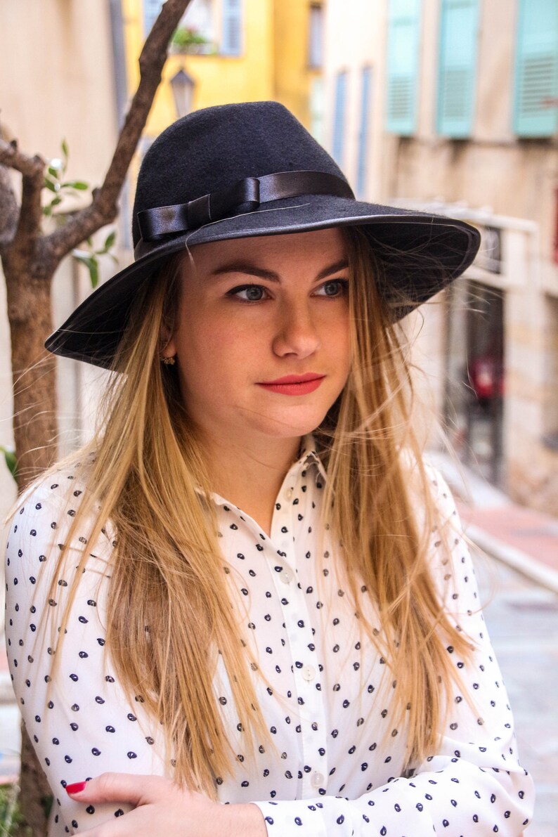 May include: A woman with long blonde hair wearing a black fedora hat with a black ribbon and a white button-down shirt with a black polka dot pattern.