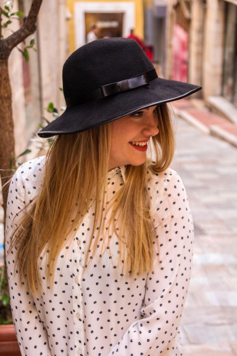 May include: A woman with long blonde hair wearing a black wide-brimmed hat with a black ribbon and a white polka dot shirt.