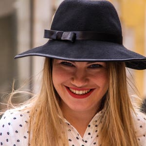 May include: A woman with long blonde hair is wearing a black wide-brimmed hat with a black ribbon and a white polka dot shirt.