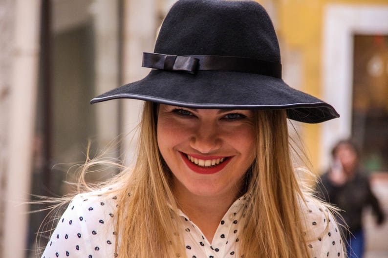 May include: A woman with long blonde hair is wearing a black wide-brimmed hat with a black ribbon and a white polka dot shirt.
