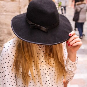 May include: A woman wearing a black wide-brimmed hat with a black bow and a white polka dot shirt.