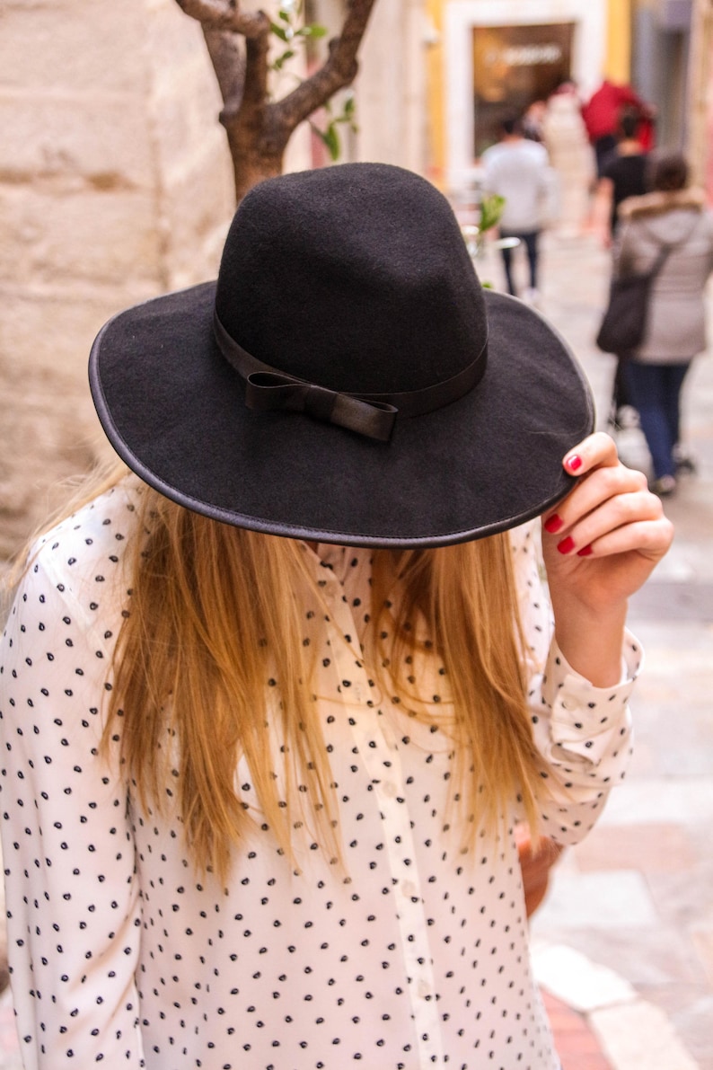 May include: A woman wearing a black wide-brimmed hat with a black bow and a white polka dot shirt.