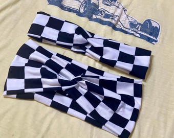 May include: Two black and white checkered headbands. The headbands are twisted at the center and are made of a soft, stretchy fabric. The background is a light yellow t-shirt with a faded blue race car graphic.