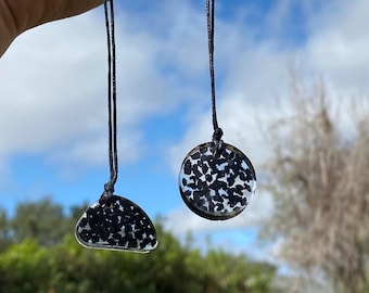 May include: Two pendants with black and white speckled designs suspended from black cords. One pendant is a rounded shape, and the other is a half-moon shape. The pendants are held up against a backdrop of a blue sky and green foliage.
