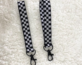 May include: Two black and white checkered wrist lanyards with black metal swivel clasps. The lanyards are made of a woven fabric with a repeating checkerboard pattern. The background is a white, fluffy surface.