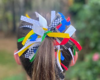 May include: A colorful hair bow featuring a variety of ribbons in white, black and white checkered, yellow, green, blue, and red. The bow is tied around a ponytail, with the back of a person visible.