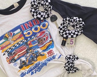 May include: A white and black raglan-style t-shirt with a graphic of the Indy 500 race from 1985, paired with black and white checkered scrunchies. The shirt features a racing car design and the text "The Greatest in Racing Spectacle".
