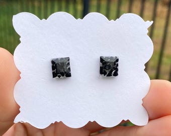 May include: A pair of square stud earrings with a black and gray speckled design. The earrings are displayed on a white, scalloped-edge card. The earrings are small, likely less than 0.5 inches in size, and appear to be made of resin or a similar material.
