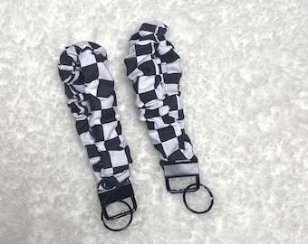 May include: Two black and white checkered wrist keychains with black metal hardware and a key ring. The keychains are laying on a white, textured surface.