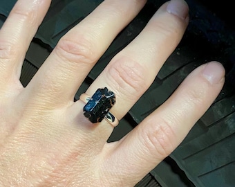 May include: A silver ring featuring a rectangular, dark blue-black gemstone. The gemstone has a rough, natural texture and is set on a simple, polished silver band. The ring is shown on a hand.
