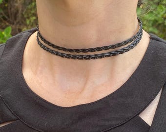 May include: A black braided choker necklace with two strands. The choker is worn around the neck and is made of a dark material. The person is wearing a black top. The background is out of focus.
