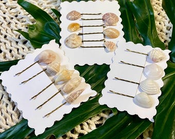 May include: Sets of seashell hair pins on white scalloped cards. The hair pins have bronze-colored metal pins and are adorned with various seashells in shades of white, tan, and pink. The background includes green leaves and a woven cream-colored surface.