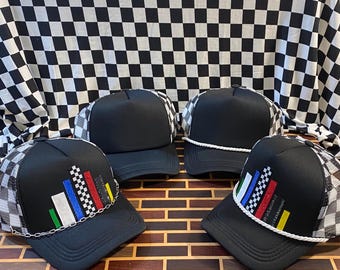 May include: Four black trucker hats with a checkered pattern on the mesh back and brim. Each hat has a black front panel and a unique design on the front, including colorful bars and chain detailing. The hats are displayed on a wooden surface.