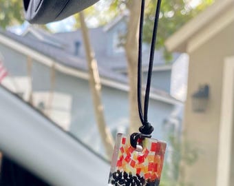 May include: A square car air freshener with a colorful mosaic design of red, orange, and black elements. The air freshener hangs from a black cord, suspended from a car's rearview mirror.
