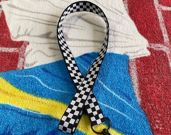 May include: A black and white checkered lanyard with a black metal clasp. The lanyard is made of a woven fabric and is designed to be worn around the neck. The background is a colorful towel.