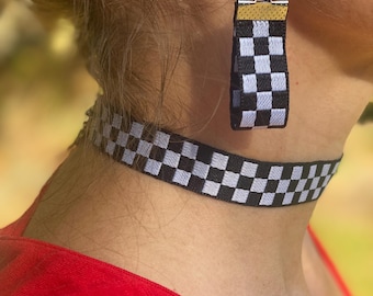 May include: A black and white checkered choker necklace and matching earring set. The earrings are rectangular with a gold-colored top. The choker and earrings have a racing flag design.