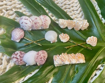May include: An assortment of seashell hair accessories, including bobby pins and barrettes. The shells are in shades of white, pink, and beige. The accessories are arranged on a large green leaf, with a white rope background.