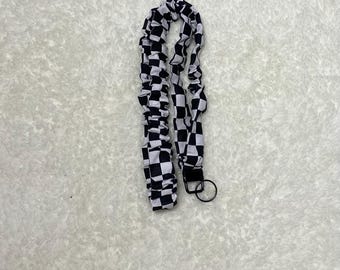 May include: A black and white checkered lanyard with a black metal clasp and key ring. The lanyard is made of a soft, fabric material and is designed to be worn around the neck.