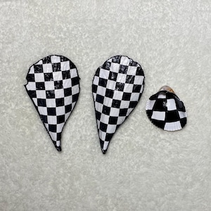 May include: Three seashells with a black and white checkered pattern. Two shells are elongated, teardrop-shaped, and the third is a smaller, round shell. The shells are arranged on a white, textured surface.