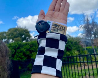 May include: Black and white checkered wrist wallet holding a car key and a ten-dollar bill. The wrist wallet is made of a soft fabric and has a zipper closure. The background is a bright blue sky with some clouds.