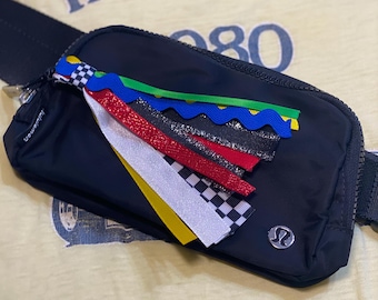 May include: Black fanny pack with a colorful ribbon tassel. The bag has a zipper closure and a silver logo. The background is a light yellow t-shirt with the words "INDY 500" printed on it.