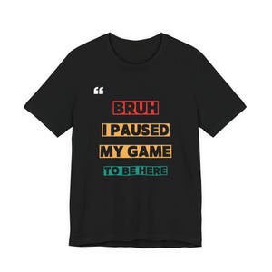 Puede incluir: Camiseta negra con el texto "BRUH I PAUSED MY GAME TO BE HERE" en rojo, amarillo y turquesa. La camiseta tiene una comilla en la parte superior izquierda.