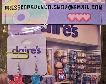 May include: A pair of gummy bear earrings, one pink and one blue, attached to a Claire's earring display card. The card is held in a plastic bag with a paperclip. The background features a store interior and the text "PRESSEDPAPERCO.SHOP@GMAIL.COM".
