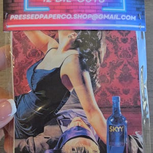 May include: A package of 12 die-cuts featuring a romantic scene with a woman in a black dress and a man with a blindfold. A blue SKYY Vodka bottle is also visible. The top of the package has text that reads "12 DIE-CUTS".