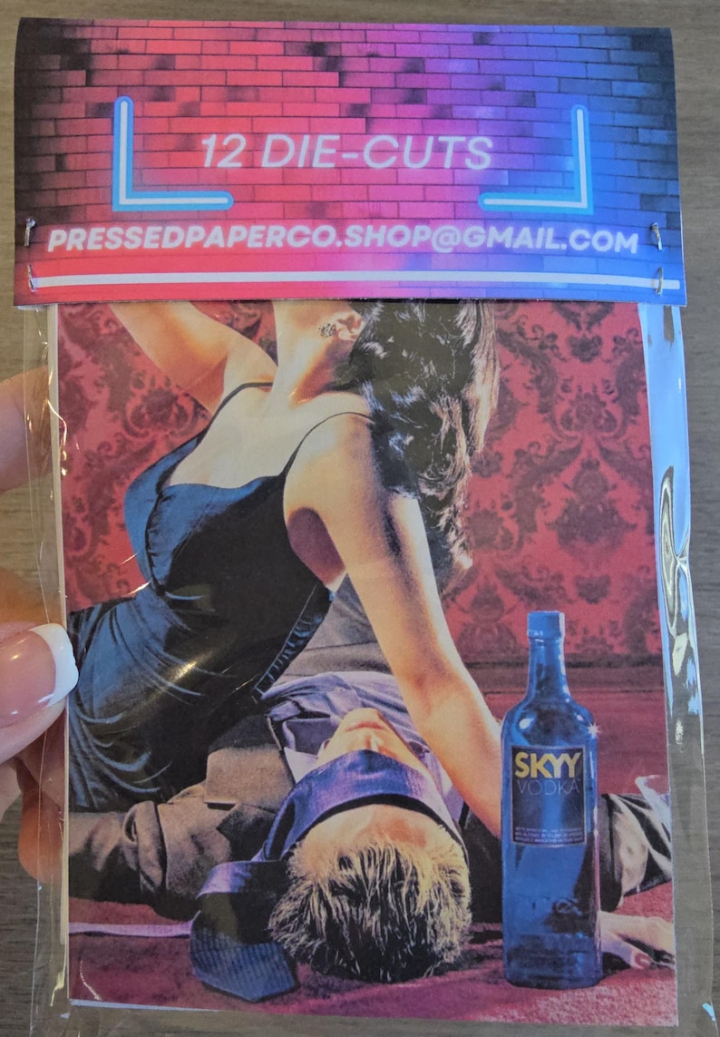 May include: A package of 12 die-cuts featuring a romantic scene with a woman in a black dress and a man with a blindfold. A blue SKYY Vodka bottle is also visible. The top of the package has text that reads "12 DIE-CUTS".