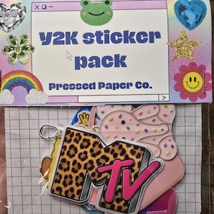 May include: A Y2K sticker pack with a variety of designs, including a leopard print MTV logo, a rainbow, and a smiling flower. The pack is packaged in a clear plastic sleeve with a grid-patterned backing.
