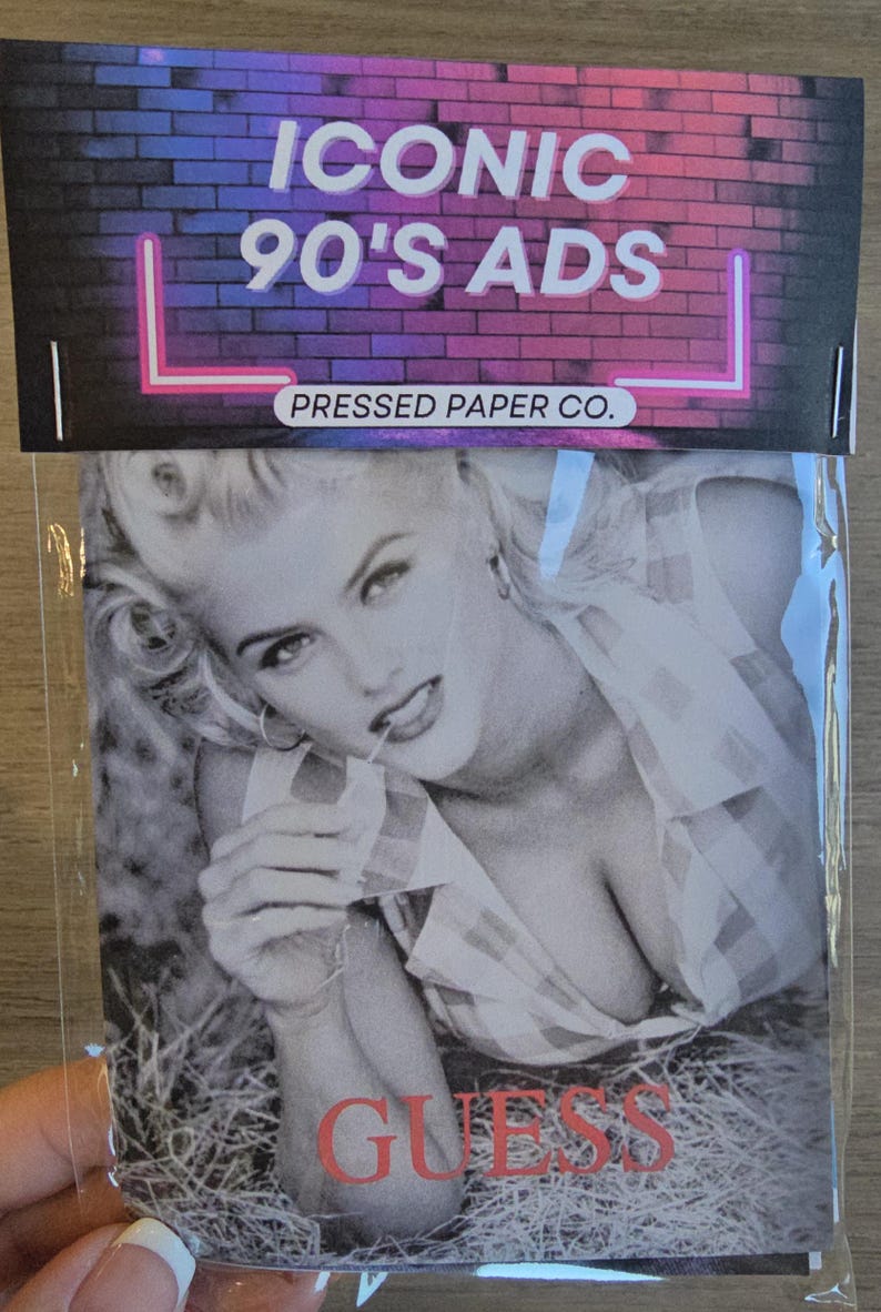 May include: A package featuring a black and white image of a woman from a Guess ad. The package has the text "ICONIC 90'S ADS" in white and pink neon against a brick background. The word "GUESS" is in red.