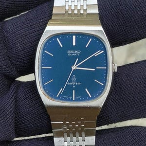 May include: A silver-tone Seiko Quartz Emblem wristwatch featuring a deep blue dial with thin silver hour markers and hands. The watch has a square-shaped case with rounded corners and a matching silver-tone metal link bracelet strap.