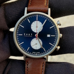 May include: A wristwatch with a dark blue face, silver casing, and a brown leather band. The watch face features white sub-dials, silver hands, and the text "Knot Timepiece". The watch also displays the date and has two side buttons.
