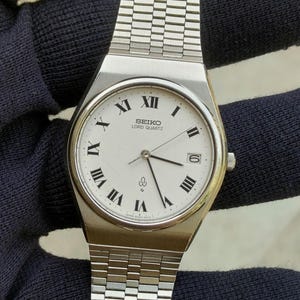 Op de afbeelding: Een zilverkleurig Seiko Lord Quartz polshorloge met een witte wijzerplaat, zwarte Romeinse cijfers, een datumvenster op de drie-uurpositie en een metalen schakelarmband. De wijzerplaat bevat de tekst Seiko Lord Quartz.