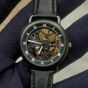 May include: A Citizen wristwatch with a black leather strap and a round black case. The dial features a skeletonized design revealing gold-toned internal gears, white Roman numeral hour markers, and white hour, minute, and second hands.