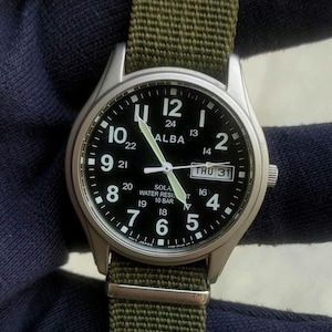 May include: An Alba solar wristwatch with a black dial, white Arabic numerals, and a day-date window at the three o'clock position. The watch features a silver-tone metal case and a textured olive green fabric strap with a metal buckle.
