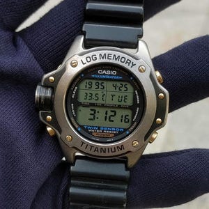 May include: A Casio Illuminator digital watch with a titanium case and black resin strap. The watch face features a multi-line LCD display showing log memory, depth, and thermo data, with text including "LOG MEMORY," "TWIN SENSOR," and "200M."