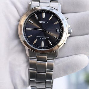 Pode incluir: Um relógio Seiko prateado com mostrador cinza escuro, com o nome da marca e o texto "Radio Wave Control Solar". O relógio tem uma janela de data às 3 horas e uma pulseira prateada. O relógio é segurado por uma mão enluvada.