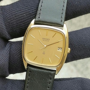 Op de afbeelding: Een goudkleurig Seiko Quartz horloge met een rechthoekige wijzerplaat en een zwarte leren band. De wijzerplaat toont de datum en het Seiko-logo. Het horloge heeft een goudkleurige bezel en een zwarte leren band.