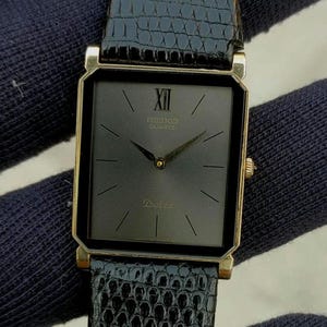 May include: A rectangular Seiko Dolce quartz watch with a black dial, gold-tone hands, and a single Roman numeral at the twelve o'clock position. The watch features a gold-tone case and a textured black leather strap with a lizard-skin pattern.