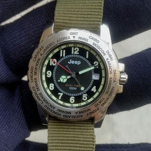 Pode incluir: Um relógio de pulso Jeep dual time com mostrador preto, algarismos arábicos brancos e janela de data. A luneta prateada apresenta nomes de cidades mundiais para rastreamento de fuso horário. Possui pulseira de nylon verde e ponteiro vermelho.