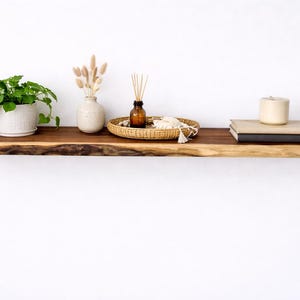 May include: A live-edge wooden shelf displays a potted plant, a small vase with dried flowers, a woven tray with a bottle and decorative items, and a stack of books with a candle. The shelf is mounted on a white wall.