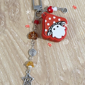 May include: A whimsical keychain with a red, polka-dotted charm featuring a ghost design. The keychain includes a silver clasp, decorative beads, a star charm, and a mushroom accent. The design has a playful, fantasy theme.