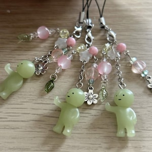 May include: Four green, glow-in-the-dark figurines with outstretched arms, attached to beaded charms. The charms feature pink, white, and clear beads, along with silver-toned flower and leaf charms. Each charm has a black cord.