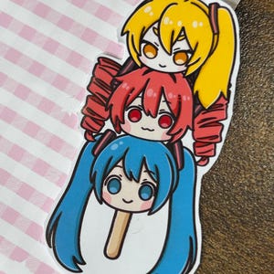 May include: A sticker featuring three anime-style characters stacked on top of each other. The top character has yellow hair, the middle character has red hair, and the bottom character has blue hair. The sticker is on a pink and white striped background.