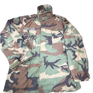 May include: Camouflage military jacket with the text "U.S. ARMY" on the front. The jacket features a high collar, multiple pockets, and a zipper closure. The pattern includes shades of green, brown, and black.